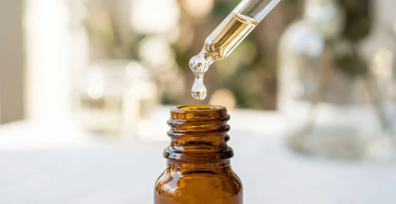 Gros plan sur une goutte d'huile essentielle tombant d'un compte-gouttes en verre dans un petit flacon ambré sur fond blanc